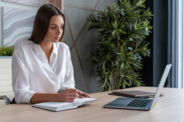 Office manager woman wearing white shirt, sitting to table with laptop and notebook, holding a pen. Concentrated office worker looking at the screen in big light office room. Concept of work