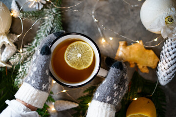 Fototapeta na wymiar childrens hands in gloves holding a cup of tea with lemon on a gray background with new year decor 