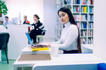 Serious adult ethnic woman looking at camera while working at laptop in library