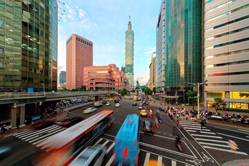 View of a busy street corner in Downtown Taipei City at rush hour with cars & buses dashing by, Taipei 101 Tower & World Trade Center Building in Xinyi Financial District & people passing on crosswalk