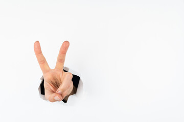 The hand shows the sign of victory and peace in close-up, the hand goes through the hole in the white wall.. Free space for text