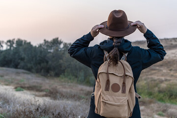 Hiker woman from back with blue jacket and backpack, holds her hipster hat, while looking at the horizon. Hike and travel concept.