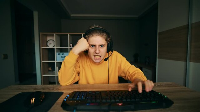 An angry gamer is sitting at a table at home and playing video games on the computer, looking at the camera with an emotionally unhappy face. Aggressive guy in headphones playing computer games