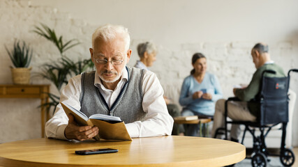 Senior man in eyeglasses reading book sitting at table in nursing home. Three elderly people, two women and disabled man, playing cards in background