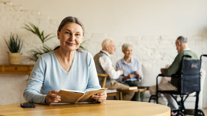 Senior woman looking at camera and smiling while reading book sitting at table in assisted living home. Three elderly people including disabled man in wheelchair playing cards in background