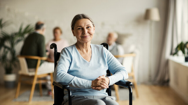Portrait of disabled senior woman in wheelchair looking at camera and smiling happily in nursing home; other aged patients in background
