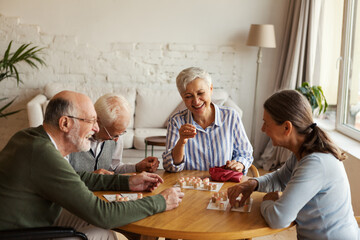 Group of four cheerful senior people, two men and two women, having fun sitting at table and playing bingo game in nursing home - obrazy, fototapety, plakaty