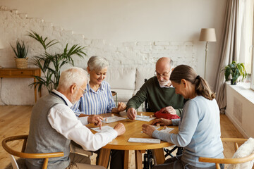 Group of four senior friends, two men and two women, sitting at table and playing bingo game in assisted living home - obrazy, fototapety, plakaty
