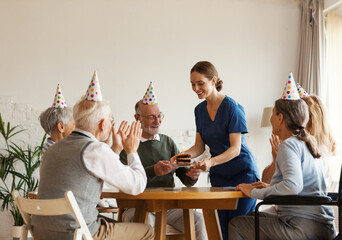 Young nurse bringing birthday cake with candle for senior man celebrating with aged friends sitting...
