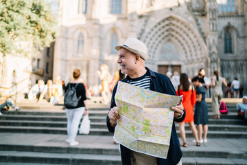 Happy Caucasian man 60s enjoying solo trip vacations for exploring Italy during retirement, cheerful male tourist in straw hat using paper orientation map for walking around historis center