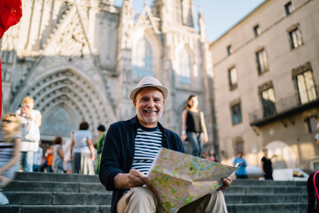 Portrait of happy male tourist with location map in hands smiling at camera during solo vacations in Spain, cheerful man in straw hat posing in historic center spending daytime for exploring town
