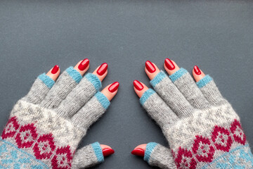 Female hands in warm winter knitted gloves with patterns with beautiful manicure - red glittered nails on gray background with copy space