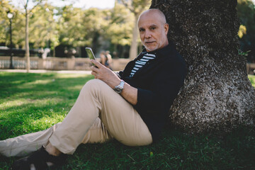 Portrait of mature male senior resting near tree in park and looking at camera during cellphone networking, aged man with modern smartphone technology in hand posing during leisure time on grass