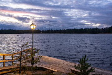 Obraz premium Finnish or russian sauna. Sunset on the lake in the evening. Wooden bath or sauna with descent into the water. Wooden flooring of planks on the shore for swimming in the cold water of the lake.