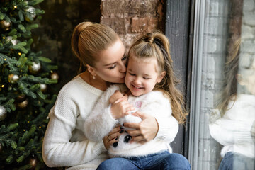 A mother is kissing in the cheek little her cute daughter near Christmas tree. New Year portrait