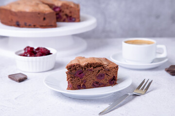 Chocolate cherry pie on a white plate. In the background is a pie, a cup of coffee, cherries and pieces of chocolate.