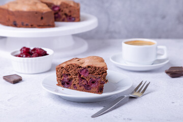 Chocolate cherry pie on a white plate. In the background is a pie, a cup of coffee, cherries and pieces of chocolate.