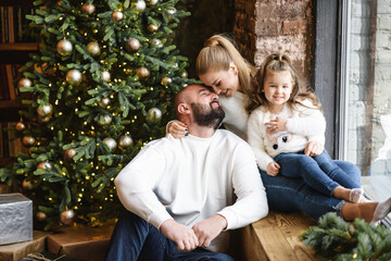 A portrait of happy family near festive Christmas tree. The young beautiful mother and little cute daughter sit on the windowsill, a bearded father sit near on the floor. Holiday with family