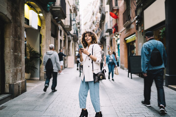 Charming female tourist in trendy clothing looking around at street architecture enjoying getaway vacations for travelling, beautiful hipster girl with modern smartphone gadget exploring city