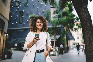 Cheerful tourist with cellular device in hand laughing at town street, joyful Caucaisan hipster girl with smartphone for travel blogging exploring urban setting during international journey