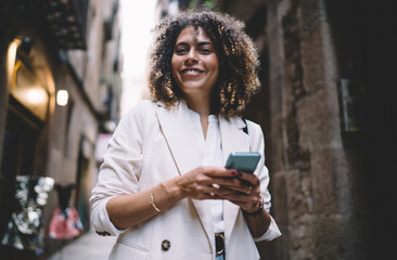 Half length of joyful hipster girl enjoying solo tourism experience posing at city street, cheerful millennial blogger with perfect curly hair holding modern smartphone gadget for networking