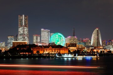 Obraz premium Night scenery of Yokohama Minatomirai Bay Area with Landmark Tower among modern skyscrapers in background, a giant Ferris wheel in an amusement park & colorful light trails of boats reflected on water