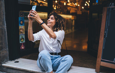 Happy Caucaisan hipster girl photographing city while resting near doorway using cellphone device for web networking, pretty female travel blogger connecting to online streams for shooting vlog