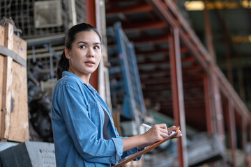 Confident Asia engineer woman worker holding a clipboard and take notes in the automotive spare parts warehouse. Looking forward to her friends. Stock management and investigation concept.