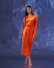 Fototapeta premium Full length studio portrait. Elegant woman standing and posing in orange silk dress against blue metaallic background. High fashion female model with make-up and wet hairstyle
