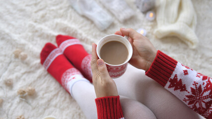 Woman hands holding a cup of hot cocoa on the background of a garland in the Christmas decorations. Cozy winter holiday concept. Woman in knitted clothes celebrating Christmas.