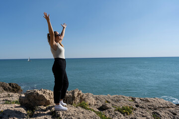 Obraz premium Pretty female celebrating and enjoying with raised up open arms standing on the rocks next to the sea.Freedom, happiness and people success lifestyle concept.Summer background.Copy space text.