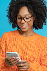 Stylish happy dark skinned millennial woman with curly hair wear orange knitted jumper, reading message from boyfriend, chatting in social media, texting sms, isolated on studio blue background. 