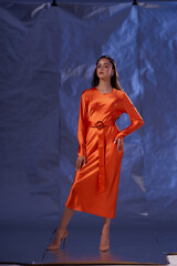 Fototapeta premium Full length studio portrait. Elegant woman standing and posing in orange silk dress against blue metaallic background. High fashion female model with make-up and wet hairstyle
