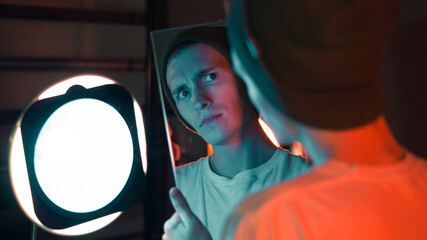 a guy holds a mirror and looks at his reflection with a surprised face, a young hipster in a hat, a red and blue neon light falls on the teenager