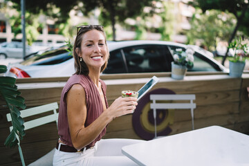Joyful woman with beverage using tablet on terrace
