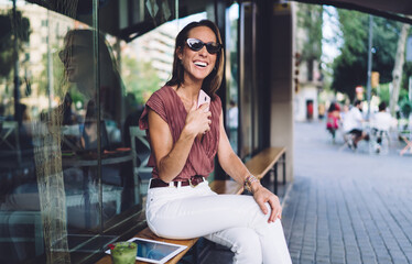 Cheerful beautiful woman in trendy wear and sunglasses sitting on cafe terrace laughing at funny social media content, smiling mature pretty female holding mobile phone waiting for call outdoors