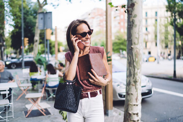 Prosperous caucasian businesswoman with notepad having working conversation on mobile phone walking on city street, successful pretty female in sunglasses talking on smartphone via roaming tariffs
