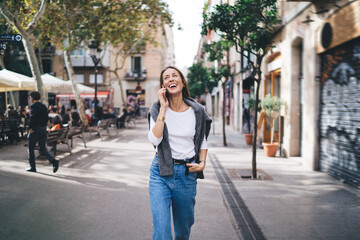 Fototapeta premium Excited female tourist in stylish apparel walking at urban street and rejoicing during positive international conversation via modern cellphone device, cheerful girl using roaming connection