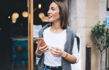 Pretty female blogger dressed in trendy wear holding modern cellphone device and looking away, charming millennial girl 30 years old using mobile device enjoying weekend and leisure time in city