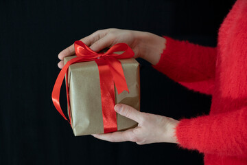 Female hands in a red sweater hold a gift wrapped in craft paper with a wide red ribbon on a black background. Holidays.