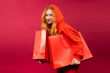 Happy young redhead girl with freckles holding red gift bags on red background.
