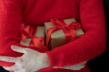 Female hands in a red sweater hold a gift wrapped in craft paper with a wide red ribbon on a black background. Holidays.