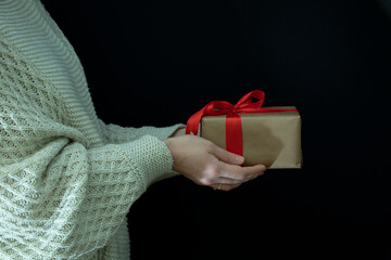Female hands in a beige knitted sweater hold a gift wrapped in craft paper with a wide red ribbon on a black background. Holidays.