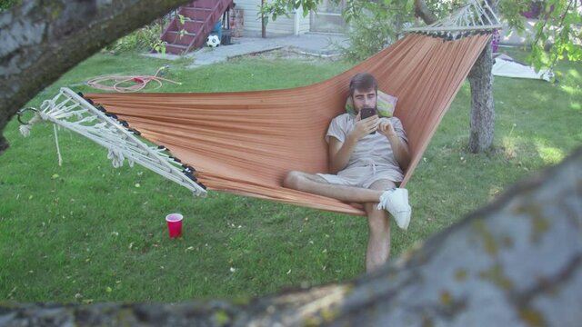 A Young Man Uses Phone While Lying In A Hammock In The Back Yard