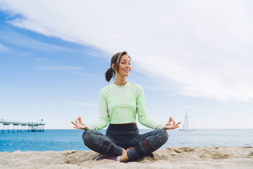 Carefree female in tracksuit sitting in lotus pose at sandy beach ready for chakra mindfulness, happy woman practice yoga exercising doing Padmasana Pranayama for morning resting at seashore