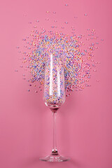 Champagne glass filled with colorful sugar balls on pink background. Glass filled with rainbow colored candies, sweet holidays concept. Celebration minimal Christmas party. Free space for text.