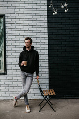 A young charismatic guy, a hipster, drinks coffee against the background of a brick wall. Dressed in a black hoodie.
