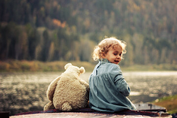 a smiling baby girl is sitting on an old boat with her teddy bear on a mountain river bank in autumn