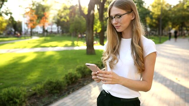 Beautiful young woman with blond hair, uses a mobile phone, exchanges messages on a smartphone, outdoors in a park