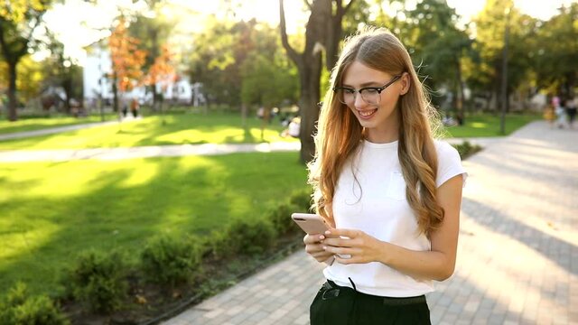 Beautiful young woman with blond hair, uses a mobile phone, exchanges messages on a smartphone, outdoors in a park